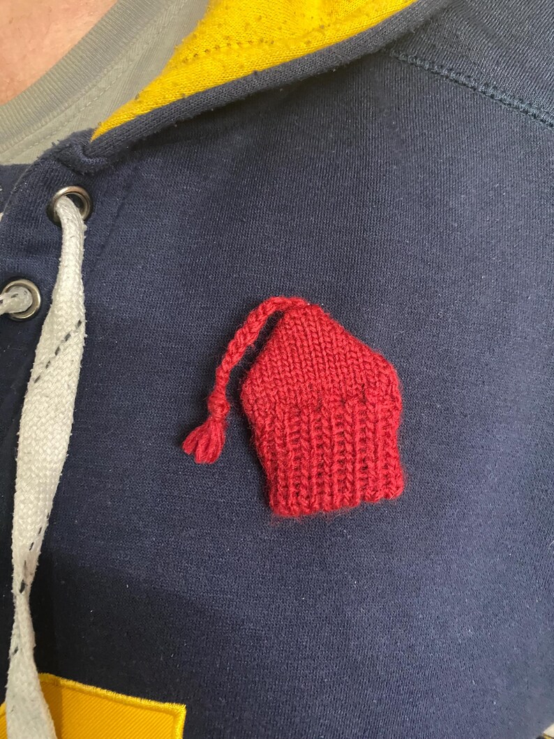 May include: A close-up of a navy blue hooded sweatshirt with a small, red, knitted hat-shaped patch. The sweatshirt has a yellow and gray collar and a white drawstring. The knitted patch is a vibrant red.