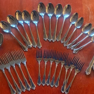 May include: A set of stainless steel cutlery, including spoons, forks, and knives, displayed on a reddish-brown surface. The cutlery features ornate handles and is likely part of a dining set.