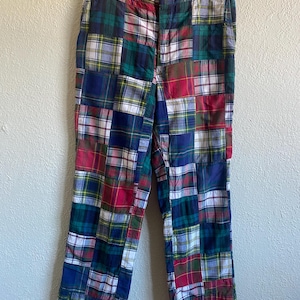 May include: Patchwork plaid pants in a variety of colors, including red, green, blue, and yellow. The pants are hanging on a hanger against a neutral background. The plaid pattern is made up of squares of different sizes.
