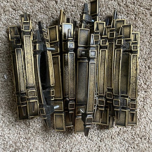 Peut inclure: Un groupe de 15 poignées de porte de placard en métal doré avec un design décoratif. Les poignées ont une forme rectangulaire avec une section centrale surélevée et un sommet pointu.