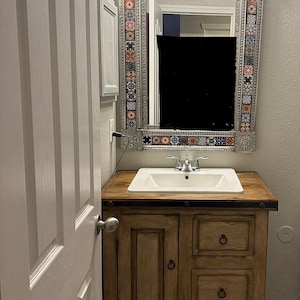May include: A bathroom vanity with a decorative mirror. The mirror has a silver frame with colorful tile accents. The vanity is made of wood and has a white sink and three drawers.