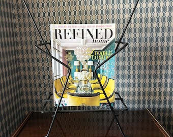 May include: A black metal magazine rack with a triangular design holds a copy of "Refined Home" magazine. The magazine features a photo of a dining room with a chandelier and yellow chairs.