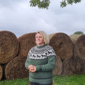 May include: A woman wearing a green sweater with a black and white horse pattern stands in front of a row of hay bales. The sweater has a crew neck and long sleeves.