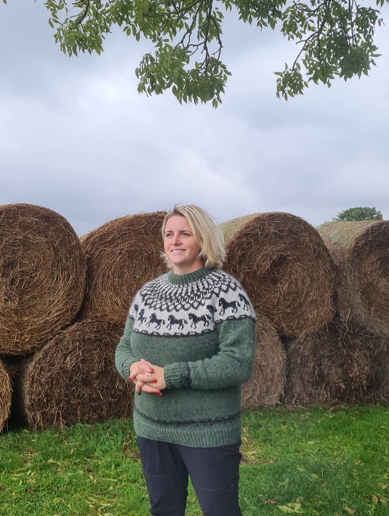 May include: A woman wearing a green sweater with a black and white horse pattern stands in front of a row of hay bales. The sweater has a crew neck and long sleeves.