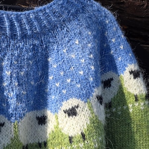 May include: A blue knitted jumper with a crew neck and a sheep design. The upper part is sky blue with white accents, transitioning to a green field with white sheep. The sheep have black faces and legs.