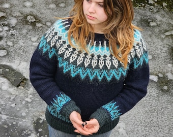 May include: A woman wearing a blue and white patterned sweater with long sleeves and a dark blue base color. She is standing on a frozen surface with ice and water.
