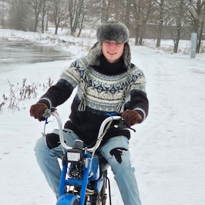 May include: A person wearing a blue and white patterned sweater and a fur hat is riding a blue moped in a snowy landscape.