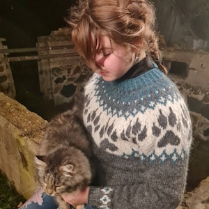 May include: A woman wearing a gray and blue sweater with a paw print pattern is holding a brown tabby cat. The sweater has a white background with blue and gray accents. The woman is wearing blue jeans with rips in the knees.