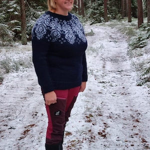 May include: A woman wearing a blue sweater with a white snowflake pattern, red pants, and black boots stands in a snowy forest.