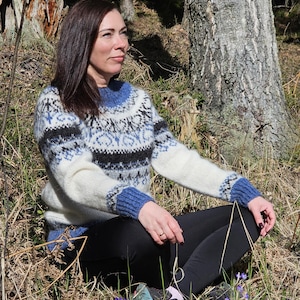 May include: A woman wearing a white, blue, and black patterned sweater sits in a forest setting. The sweater has a traditional Scandinavian design.
