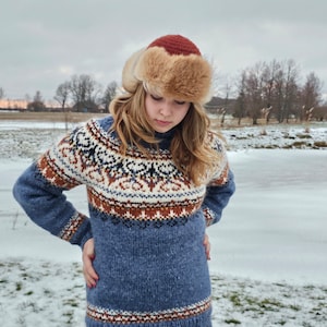 May include: A woman wearing a blue knitted sweater with a traditional Norwegian pattern, a brown fur hat with a red pompom, and brown pants. She is standing in a snowy field.