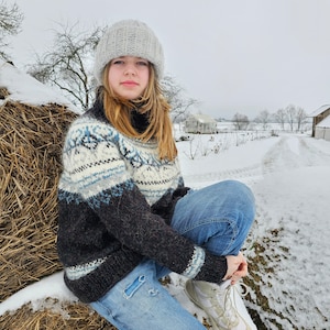 May include: A young person wearing a white knit hat, a brown and blue patterned sweater, and blue jeans sits on a bale of hay in a snowy field.