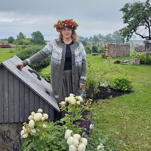 May include: A woman wearing a gray knitted cardigan with a white and blue patterned design, a red flower crown, and a gray skirt. She is standing in front of a wooden well with a green lawn and white flowers in the foreground.