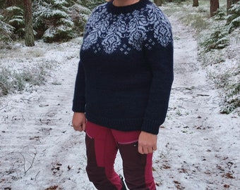 May include: A woman wearing a dark blue sweater with a white and gray patterned design, burgundy pants, and black boots. She is standing in a snowy forest.