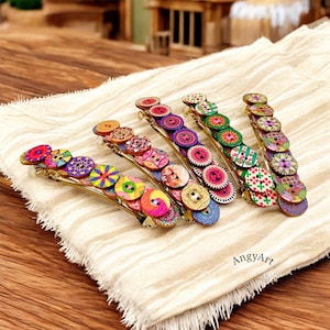Puede incluir: Cinco horquillas decorativas para el cabello con botones de madera coloridos y estampados. Los botones presentan varios diseños, incluyendo patrones florales y geométricos. Las horquillas son doradas y descansan sobre una tela color crema.