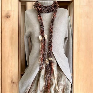 May include: A long, brown and rust-coloured scarf with a textured, looped design. The scarf is draped over a grey sweater and dress, and features cream-coloured ribbon accents. The scarf is displayed on a mannequin.