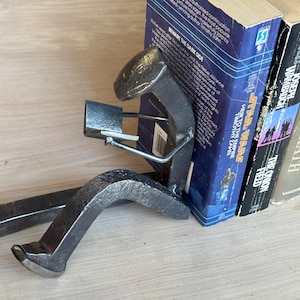 May include: A metal bookend shaped like a person reading a book. The bookend is holding a book with one hand and has a book under the other arm. The bookend is sitting on top of a stack of books. The books have titles like "Star Wars" and "Grapes of Wrath".