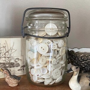 May include: A glass jar with a wire bail closure filled with white buttons. The jar has the text "Ball Store" on the side.