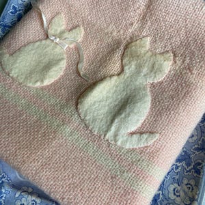 May include: A pink and white baby blanket with two white felt cat appliqués. The blanket has a striped design and is folded on a blue floral patterned plate.
