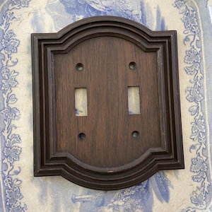 May include: A dark brown wooden double light switch cover with a decorative, layered frame. It features two rectangular switch openings and four small mounting holes. The background is a blue and white patterned surface.
