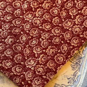 Könnte beinhalten: Ein gefaltetes Stück Stoff mit einem sich wiederholenden Blumenmuster. Der Stoff ist tiefrot mit einem cremefarbenen Blumendesign. Die Blumen sind klein und detailliert und schaffen eine Vintage-Ästhetik. Der Stoff liegt auf einem blau-weiß gemusterten Teller.