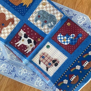 May include: A blue patchwork quilt featuring farm animals like a cow, sheep, and elephant. The quilt has a blue border with a rainbow and ark design. The quilt is made of fabric and has button details.