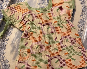 May include: A vintage doll dress with a colorful pattern. The dress features a square neckline and short sleeves trimmed with green fabric. The fabric has a grid pattern with circles and rectangles in orange, green, and purple.