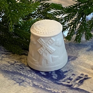 May include: A white ceramic thimble with a windmill design. The thimble has a rounded top and a flared base. The windmill is depicted with an X-shaped sail. The thimble is set against a blue and white patterned surface with green foliage.