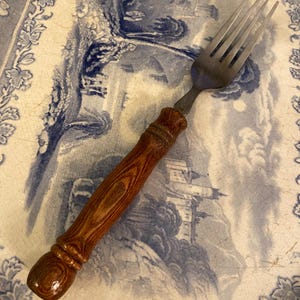 May include: A stainless steel fork with a wooden handle. The handle is a dark brown colour with a wood grain pattern. The fork is resting on a blue and white patterned plate.