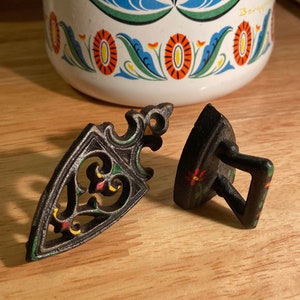 Op de afbeelding: Twee miniatuur strijkijzers van gietijzer met beschilderde bloemdetails. Een strijkijzer heeft een traditionele vorm met een handvat, de andere is een meer sierlijk ontwerp met een puntige punt.