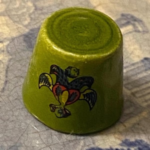 May include: A green ceramic thimble with a decorative design featuring a bird-like motif in black, red, and yellow. The thimble is a sewing tool, designed to protect the finger while pushing a needle through fabric.