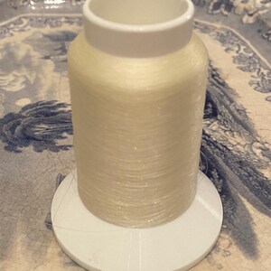 May include: A spool of white thread on a white plastic bobbin. The thread is wound tightly and has a slight sheen.