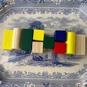 May include: A selection of brightly coloured wooden blocks in different shapes, including rectangles, squares, and cylinders. The blocks are painted in vibrant colours such as yellow, green, red, and blue, and are arranged on a decorative blue and white plate.
