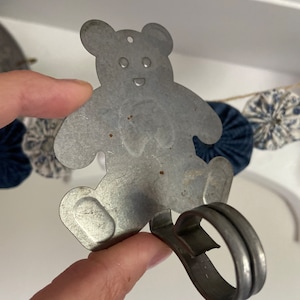 May include: A silver metal teddy bear hook with a rounded hook at the bottom. The hook is designed to hang items on a wall or door.
