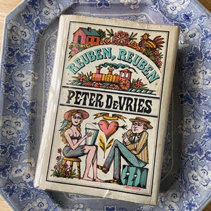 May include: A vintage book titled "Reuben, Reuben" by Peter DeVries, featuring a colorful illustration of a train, house, and figures. The book is on a blue and white floral patterned plate.