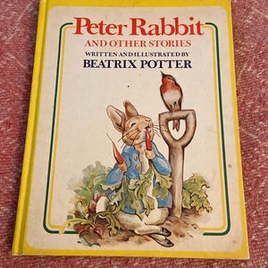 May include: A yellow book cover with a picture of Peter Rabbit eating a carrot. The book title is "Peter Rabbit and Other Stories" written and illustrated by Beatrix Potter. A robin is perched on a shovel handle.