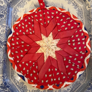 May include: A circular quilted pot holder featuring a red and white paisley pattern and a central star design. The star is made of red fabric and a floral print. The edge has a red, white, and blue trim, and a hanging loop.