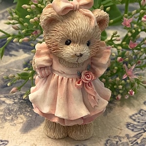 May include: A detailed figurine of a teddy bear in a pink dress and bow. The bear has a light brown fur texture and stands on a patterned surface. A pink rose adorns the dress. The background includes greenery and small pink flowers, creating a delicate scene.