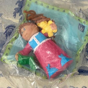 May include: A plastic Cabbage Patch Kid doll wearing a pink and blue nightgown, holding a yellow teddy bear and a green blanket. The doll is packaged in a clear plastic bag with a green and blue background.