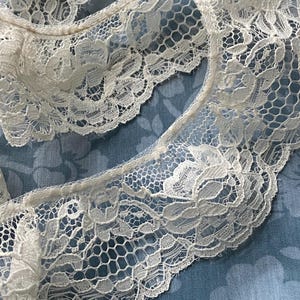 May include: Close-up of cream-coloured lace trim featuring a floral design. The lace has a scalloped edge and an open weave. The background is a blue fabric with a subtle floral pattern.