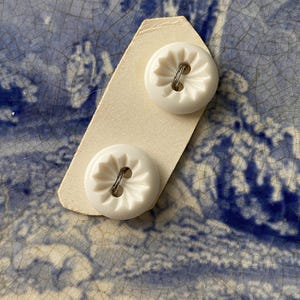 Peut inclure: Deux boutons ronds blancs avec un motif en forme d'éclat d'étoile sont attachés à une carte de couleur crème. Chaque bouton a une boucle métallique pour la couture. Les boutons sont posés sur un fond à motifs bleu et blanc.