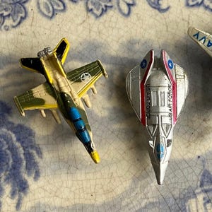 May include: Two toy airplanes. One is a camouflage green and tan jet with a yellow nose and black accents. The other is a silver jet with red and blue accents and the text "US AIR FORCE".
