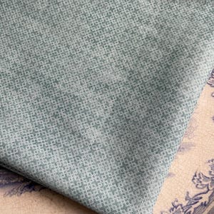 May include: Close-up of a folded piece of fabric with a small, repeating pattern in shades of green and white. The fabric has a textured appearance. The fabric is resting on a decorative plate with blue floral designs.