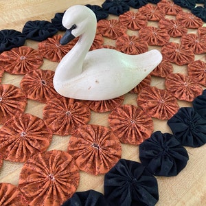 May include: A white wooden swan figurine sits on a handmade table runner. The runner is made of fabric circles in black and orange with a floral pattern.