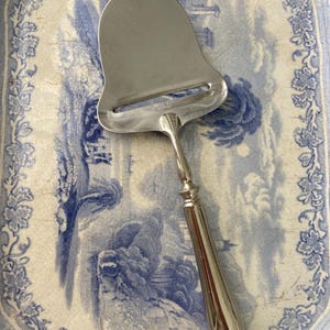May include: A silver cheese slicer with a curved blade and a decorative handle rests on a blue and white patterned plate. The plate features a landscape design and floral border. The cheese slicer is a kitchen utensil.