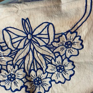 May include: A close-up of a cream-colored fabric with a blue embroidered design. The design features a large bow and several stylized flowers.