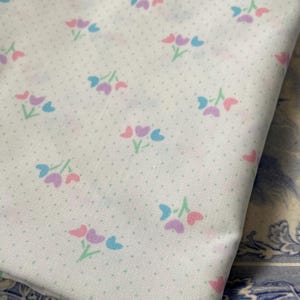 May include: White fabric with a repeating pattern of small, pastel-colored floral clusters. Each cluster features pink, blue, and purple tulip-like shapes with green stems. The fabric has a subtle polka dot texture.