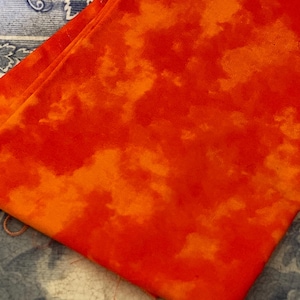 May include: A folded piece of fabric with a vibrant orange and red cloud-like pattern. The fabric is layered over a blue and white patterned surface, creating a striking contrast. The material appears soft and possibly suitable for apparel or home decor.