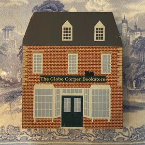 May include: A wooden decorative piece shaped like a two-story building with a dark gray roof and red brick facade. The building has white-framed windows and a green door. The text "The Globe Corner Bookstore" is above the windows.