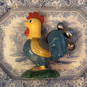 May include: A ceramic rooster figurine with a yellow body, red comb, and blue tail. The rooster is on a green base and sits on a blue and white patterned plate. Silver measuring spoons are next to the rooster.